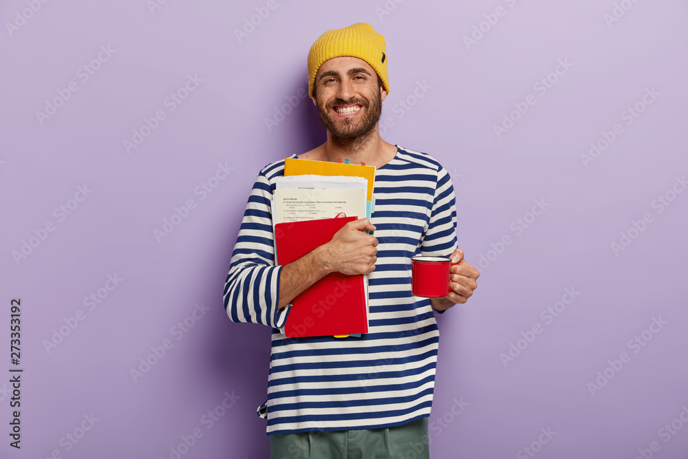 Happy college student carries papers and notepad, prepares for exams, drinks hot beverage from cup, wears striped clothing, isolated over purple wall, studies at high school. Education concept Happy college student carries papers and notepad, prepares for exams, drinks hot beverage from cup, wears striped clothing, isolated over purple wall, studies at high school. Education concept