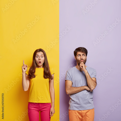 Shocked woman points upwards, keeps mouth opened, thoughtful unshaven man holds chin, stand shoulder to shoulder, pose over purple and yellow background, free space for your promotional content
