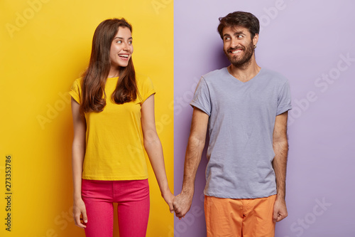 Romantic couple in love have date, hold hands, look positively at each other, feel support, walk together. Positive man poses over purple background, woman on yellow. Contrast. Relationship concept photo