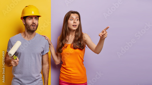 Home renovation, painting and decoration concept. Shocked husband wears helmet, holds paint roller, poses with wife, busy painting walls in room at purple and yellow color, redecorate apartment