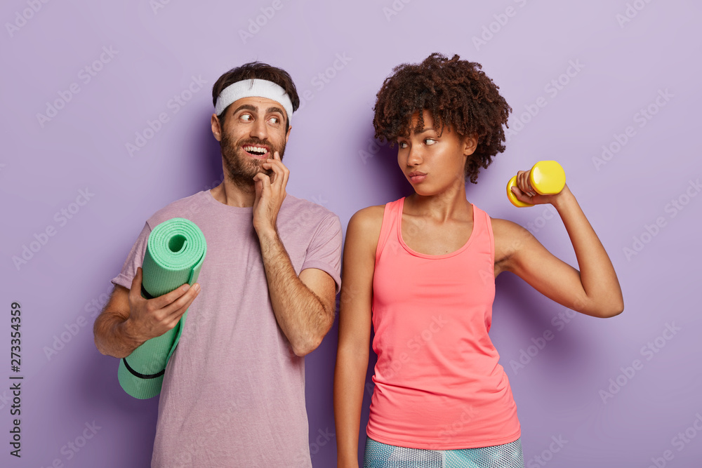 Fototapeta premium Indoor shot of happy multiethnic couple have early morning workout, do sport exercises with dumbbell, going to have yoga training on fitness mat, stands closely. Cheerful sporty man and serious woman
