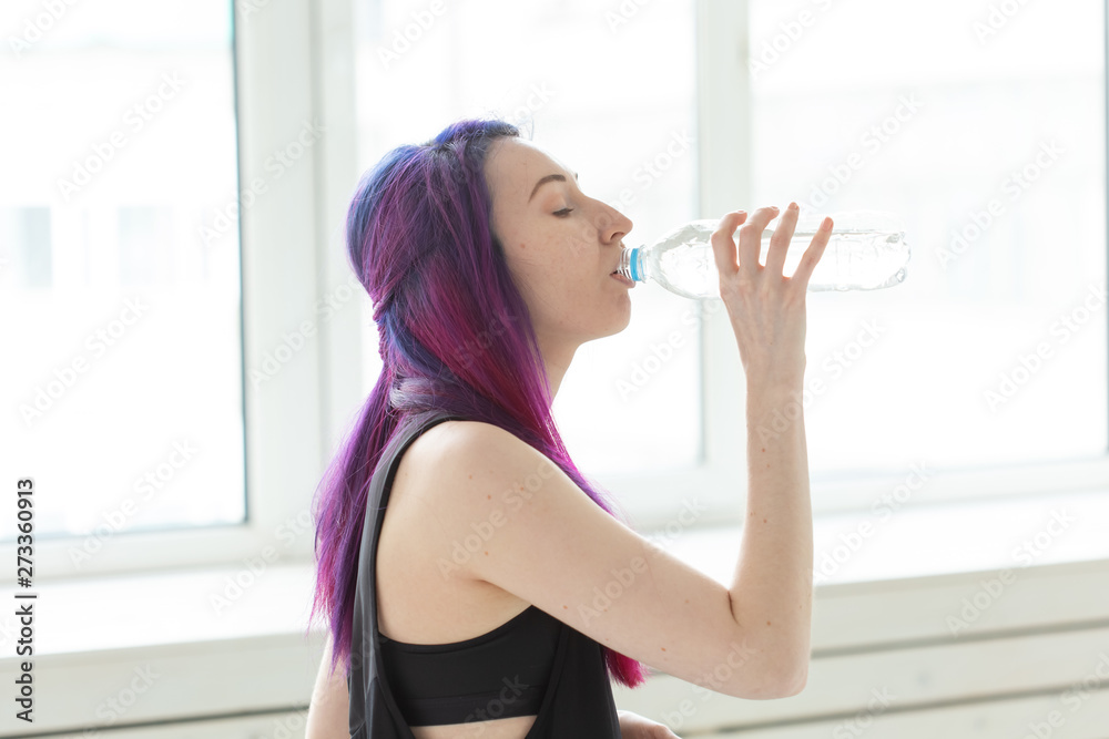Young girl hipster with colored hair drinks water from a bottle after intense physical training in the gym. The concept of thirst and water balance of the body. Young girl hipster with colored hair drinks water from a bottle after intense physical training in the gym. The concept of thirst and water balance of the body.