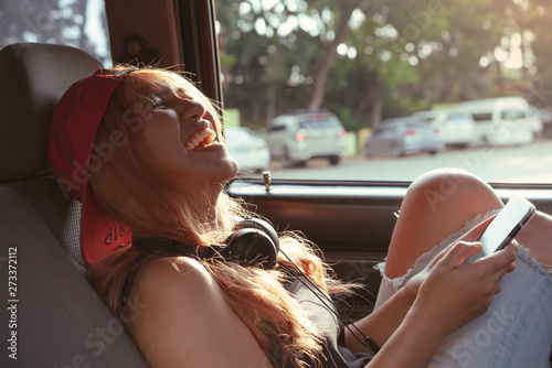 Cool Asian hipster girl laughing & messaging on phone inside car road trip