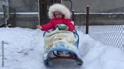 Baby girl is sitting on a sleigh hiding in a blanket, catching the flying snowflakes and reading a book