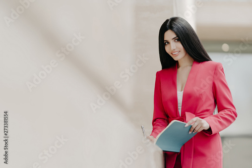 Beautiful smiling lady in elegant red costume, writes some notes in notepad, stands outside, copy space on left side for your promotional content. Businesswoman has meeting with partners or colleagues