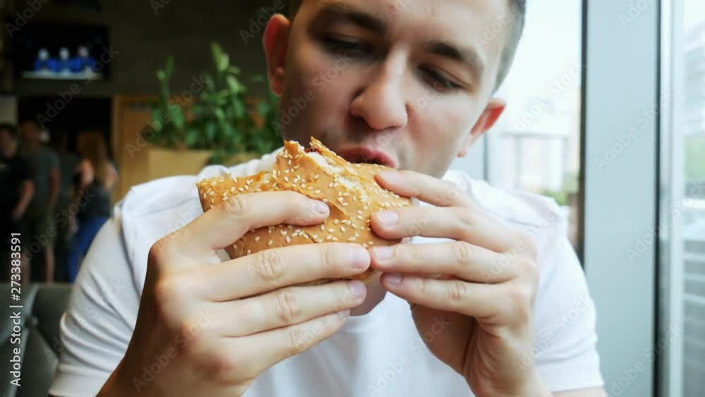 Close-up portrait of young hungry guy with pleasure eats hamburger in a cafe, sharpness on a hamburger, fast food restaurant Close-up portrait of young hungry guy with pleasure eats hamburger in a cafe, sharpness on a hamburger, fast food restaurant