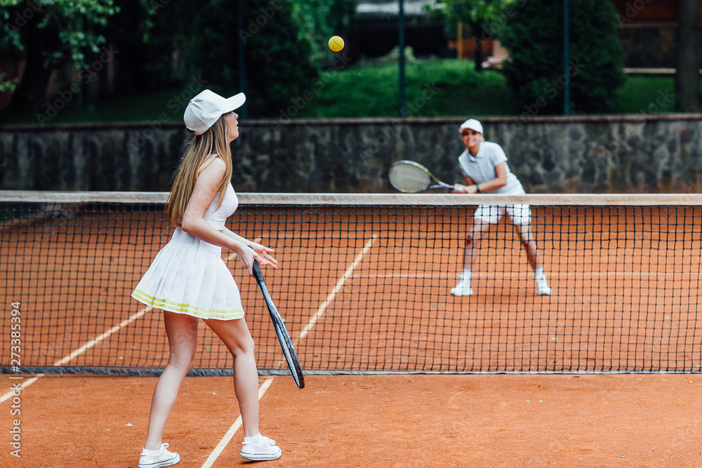 Two fit woman in cap and tennis uniform serving tennis ball during training on the outdoor tennis court.