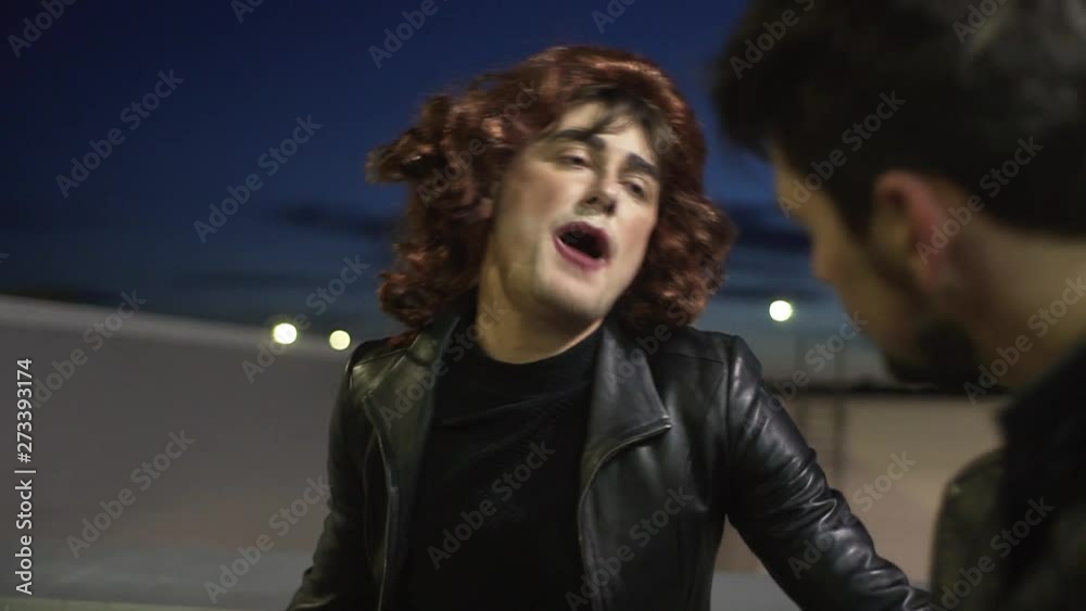 Comic young male dressed like woman with make-up wearing black tight leather jacket with blouser and brown curly wig, emotionally sings standing outside next to other brunette man. Comic young male dressed like woman with make-up wearing black tight leather jacket with blouser and brown curly wig, emotionally sings standing outside next to other brunette man.