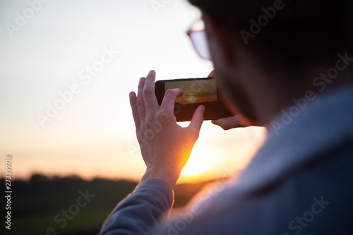Back view of young guy take pictures of sunset by smartphone