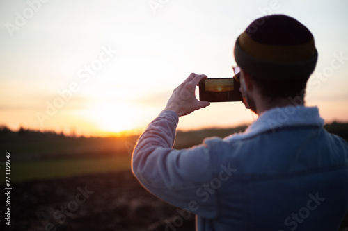 Back view of young guy take pictures of sunset by smartphone