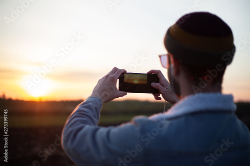 Back view of young guy take pictures of sunset by smartphone