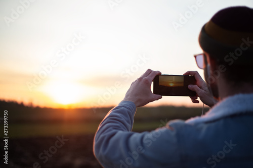 Back view of young guy take pictures of sunset by smartphone