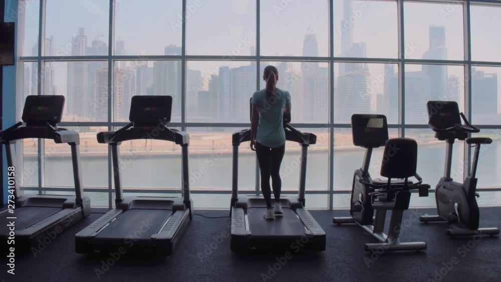 Woman running on treadmill at a panoramic window with city view at the sunrise. Woman running on treadmill at a panoramic window with city view at the sunrise.