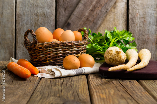 Fototapeta Naklejka Na Ścianę i Meble -  basket of eggs and carrots, parsley roots, celery, cutting board, old weathered wooden background