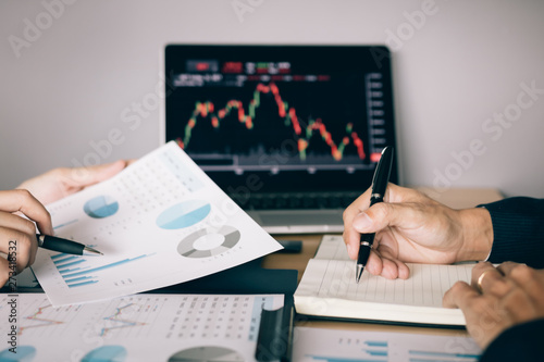 Two investors are working together with analyzing the stock data graphs in the paper and viewing the data on the laptop screen.