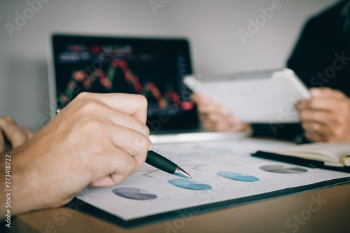 Two investors are working together with analyzing the stock data graphs in the paper and viewing the data on the laptop screen.