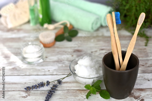 Three eco friendly wooden toothbrushes in brown ceramic glass. Coconut oil in glass jar with green mint leaves, lavender flowers in the blurred background. Natural zero waste concept.Selective focus.