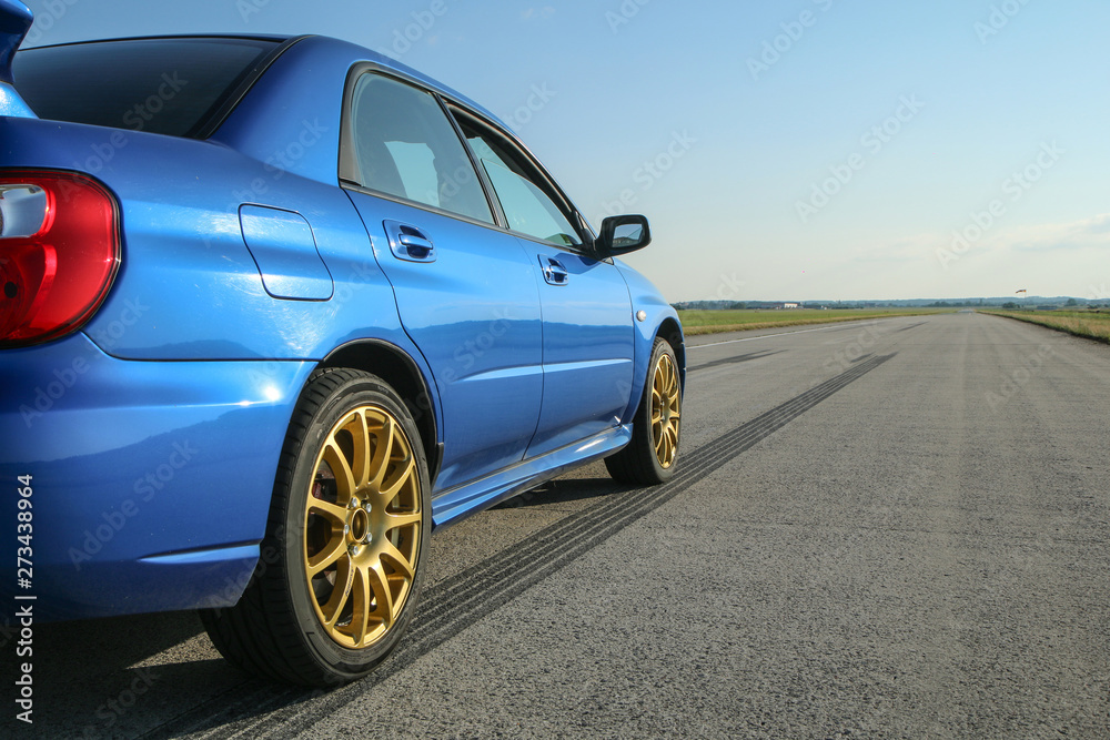 The blue youngtimer sports car with golden rims is standing on the concrete runway of the airport and is ready to drive fast. Like being part of the drag race. The rubber tracks are visible. 