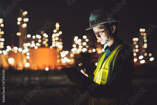 Asian man engineer using digital tablet working late night shift at petroleum oil refinery in industrial estate. Chemical engineering, fuel and power generation, petrochemical factory industry concept