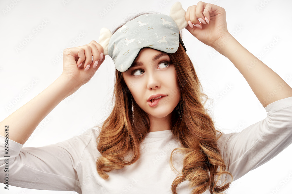 Thoughtful relaxed young woman has curly hair, wears eyemask, slumber suit, looks away intently and bites her lips. Beautiful lady model is standing at white background in Studio Thoughtful relaxed young woman has curly hair, wears eyemask, slumber suit, looks away intently and bites her lips. Beautiful lady model is standing at white background in Studio