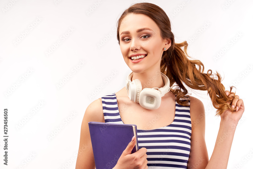 A long-haired curly-haired girl with headphones on her neck holds a notebook in her hand and cheerfully smiles looking at the camera. Studio portrait on white background A long-haired curly-haired girl with headphones on her neck holds a notebook in her hand and cheerfully smiles looking at the camera. Studio portrait on white background