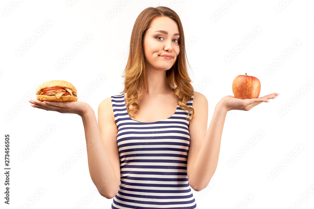 young woman holding Apple and Burger, on her face dissatisfied and irritated emotions from the choice between healthy food and delicious fast food. The concept of healthy eating and weight control