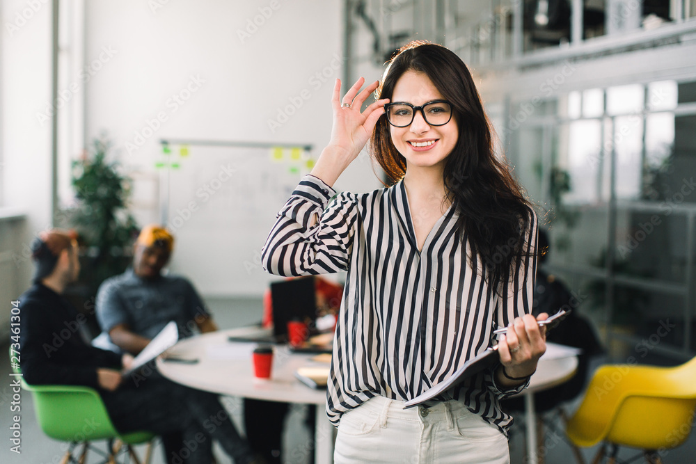 Beautiful trendy brunette business woman in glasses with folder of documents in her hands indoors. Team of young workers discussing on the background