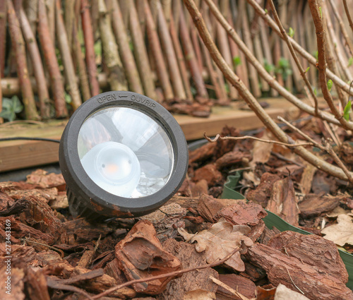 Closeup of a LED graden lamp, stuck in a flowerbed and between bark mulch of pine trees. Bulb of the modern lamp can be seen through the glass front. Lamp is powered by a solar module.