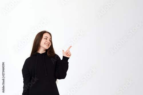 Portrait of a pretty young brunette in a black sweatshirt on a white background pointing at the right side with fingers and looking at the camera.