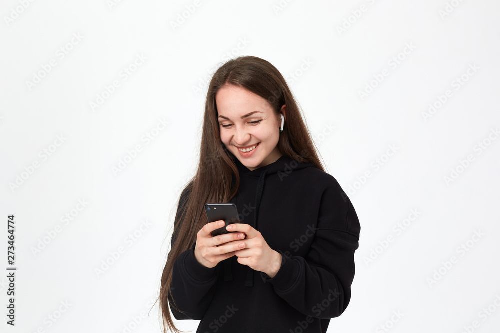 Studio shot of a beautiful young brunette woman with wireless headset looking happy at the phone while standing over white background. Studio shot of a beautiful young brunette woman with wireless headset looking happy at the phone while standing over white background.