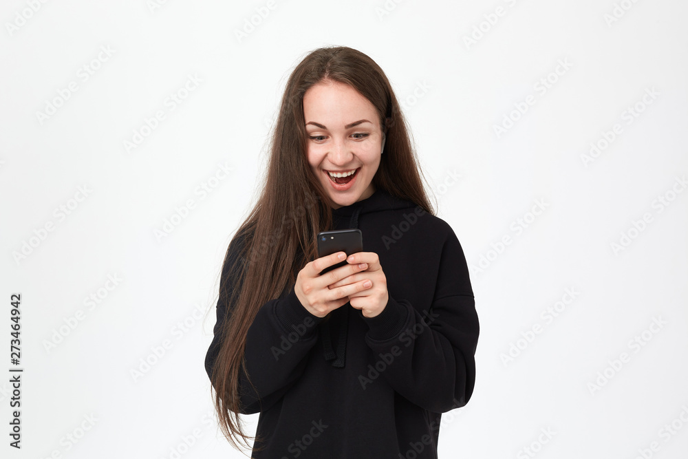 Studio shot of a beautiful young brunette woman with wireless headset looking happy at the phone while standing over white background. Studio shot of a beautiful young brunette woman with wireless headset looking happy at the phone while standing over white background.