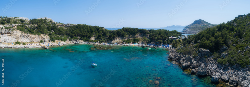 Sunny beach lagoon with rocky coastline, Greece. Tourists under umbrella chill relax near clear blue emerald turquoise sea water