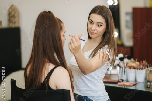 Beautiful femalemakeup artist doing makeup for a young redhead girl in a beauty salon sitting in front of a large mirror. Concept of preparation for the holiday and meeting