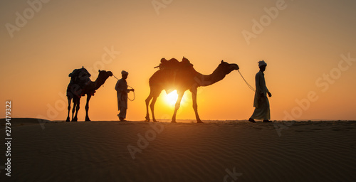 Rajasthan travel background - Two indian cameleers (camel drivers) with camels silhouettes in dunes of Thar desert on sunset. Jaisalmer, Rajasthan, India