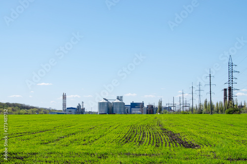 Modern soybean processing plant, agricultural Silos against green field and blue sky. Storage and drying of grains, wheat, corn, soy, sunflower. Agricultural silo at feed mill factory