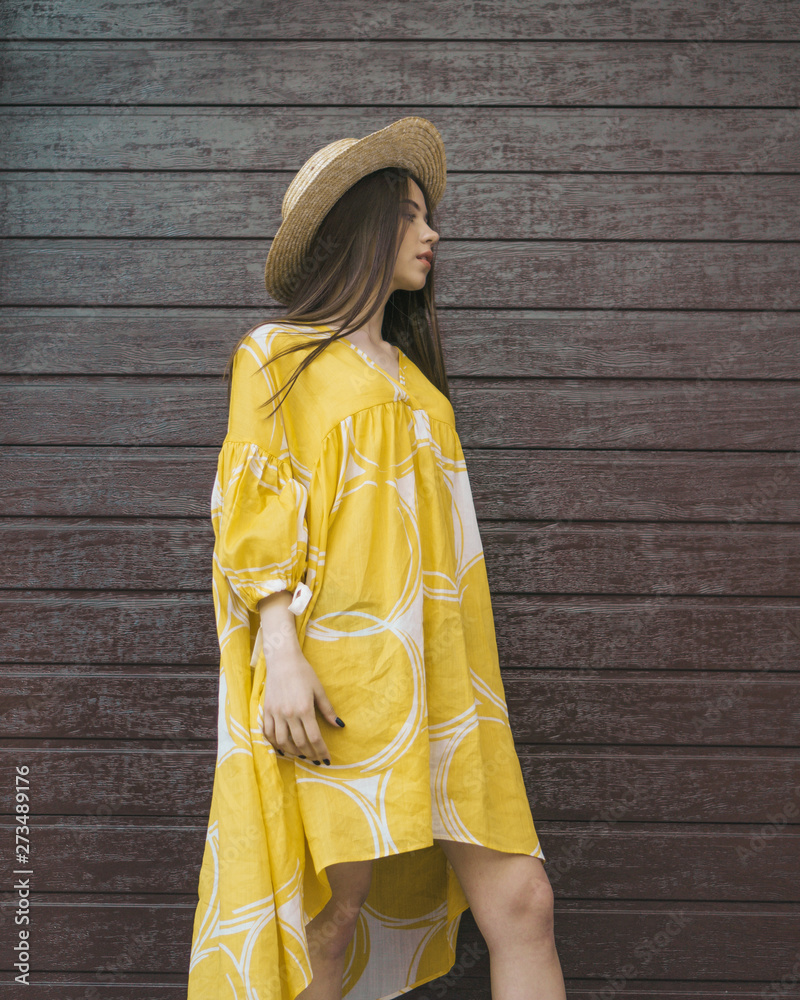 Fototapeta premium Beautiful girl model posing on the background of brown wall in summer yellow dress.