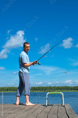 a man in yellow glasses on the pier, throws a fishing rod for fishing, against the blue sky and the lake