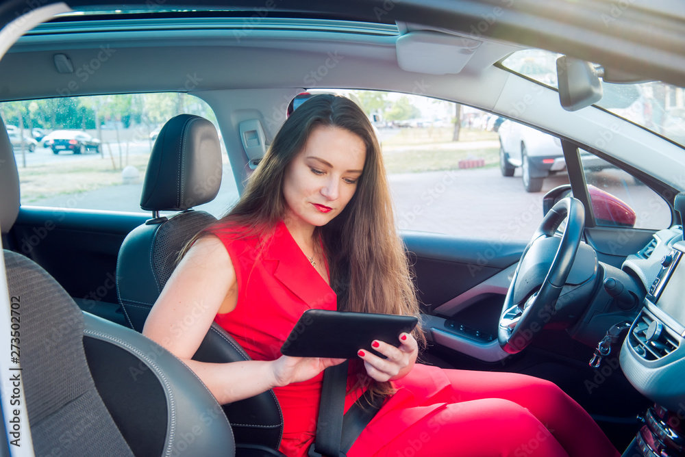 Portrait of serious business lady, caucasian young woman driver in red summer suit setting up the route on a navigator while sitting behind the wheel car. Side view. Selective focus, copy space.