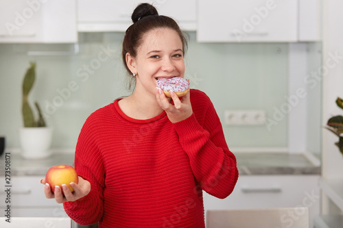 Pregnant woman eating sweet cake and holding apple in another hand, stands wearing red casual sweater in front white kitchen interior, female expresses happyness and gladness. Pragnancy concept. photo