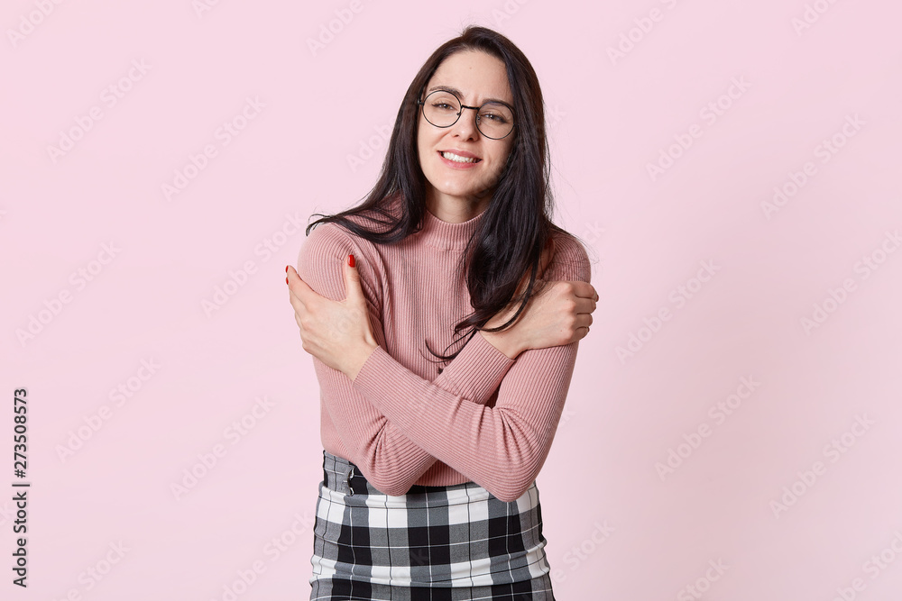 Closeup portrait of attractive good looking female, hugging herself on account of cold, wearing pink sweater, skirt and stylish eyeglasses, trembling, setting teeth, isolated over pink background.