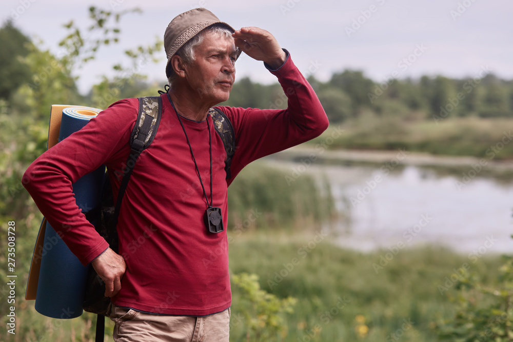 Obraz premium Photo of caucasian man hipster man with backpack and rug in nature background. Relax time on holiday, eldery male looking far away, dressed casual red shirt and cap. Traveling and active rest concept.