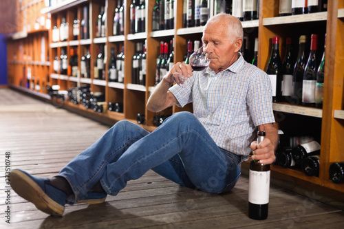 Portrait of senior man sitting on floor in winery tasting room, drinking red wine
