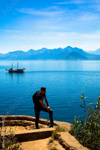 Young man stands on a rock on the background of a beautiful view of the Mediterranean Sea, the mountains and the ship. Turkey, Antalya.