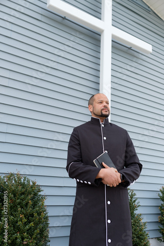 A african-american Male Reverend stands in front of a church with a bible and a black robe