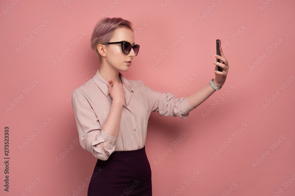 Pink-haired young woman with sunglasses takes a selfie on her smartphone, pink background Pink-haired young woman with sunglasses takes a selfie on her smartphone, pink background