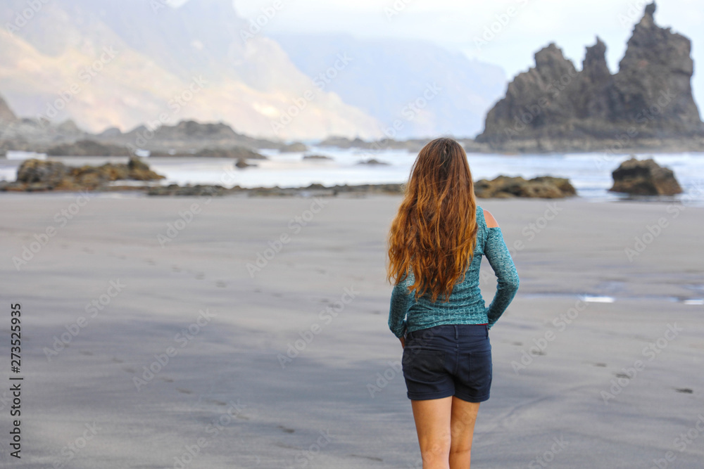 Back view of girl walking reflexive e tranquil on hidden amazing black beach on sunrise. Young female discovering wild paradisiac beach in Tenerife Island.