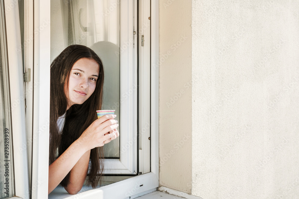 young brunette hipster girl is looking out of open window of house, holding a colored paper disposable cup with coffee or tea in her hands, using disposable eco-friendly tableware young brunette hipster girl is looking out of open window of house, holding a colored paper disposable cup with coffee or tea in her hands, using disposable eco-friendly tableware