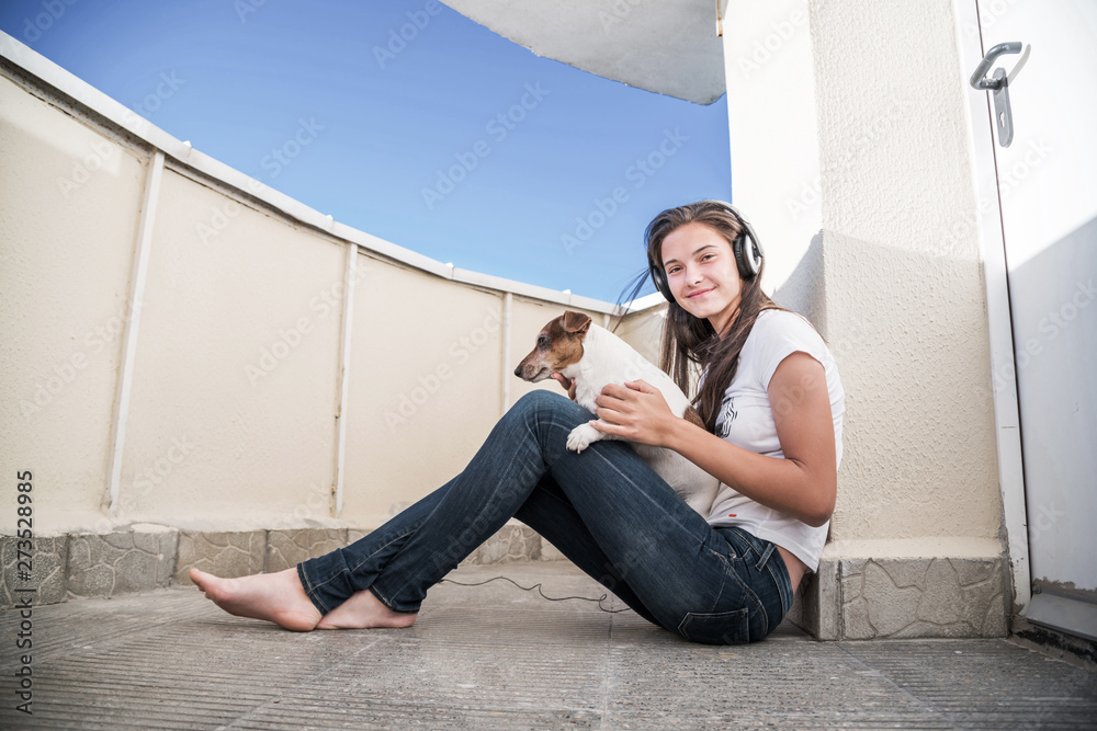 barefoot hipster girl in jeans is sitting on balcony in headphones with pet  dog in her arms, listening to music and smiling Stock Photo | Adobe Stock