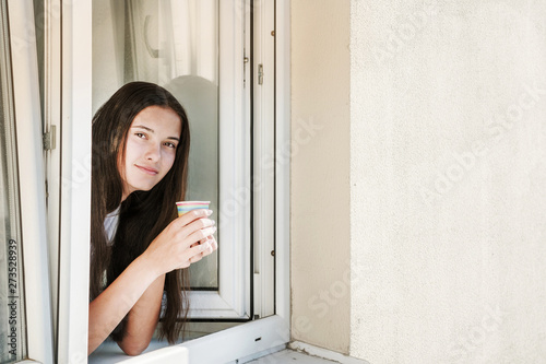 young brunette hipster girl is looking out of open window of house, holding a colored paper disposable cup with coffee or tea in her hands, using disposable eco-friendly tableware