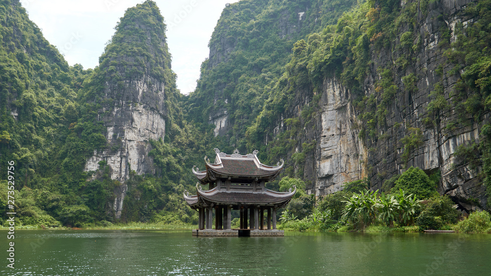 Small temple on the river surrounded by karst mountains. Beautiful landscape during the Trang An boat tour in Tam Coc, Vietnam.